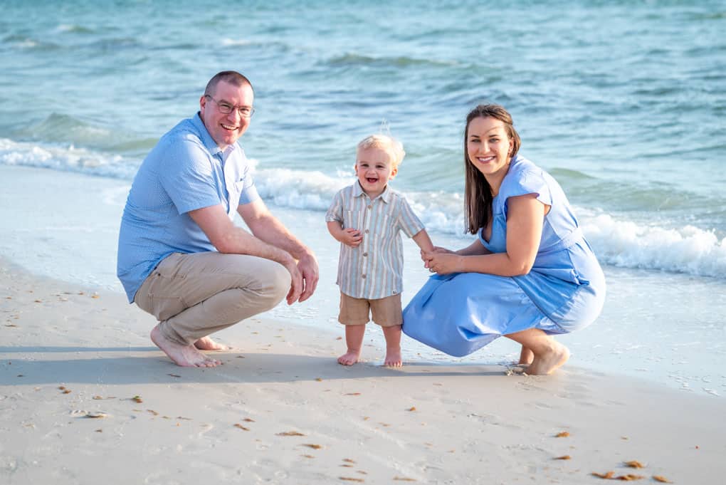 A smiling family of three, with a man, woman, and young child, kneel and stand on a sandy beach near gentle ocean waves under a sunny sky, captured by a Naples beach portrait photographer.