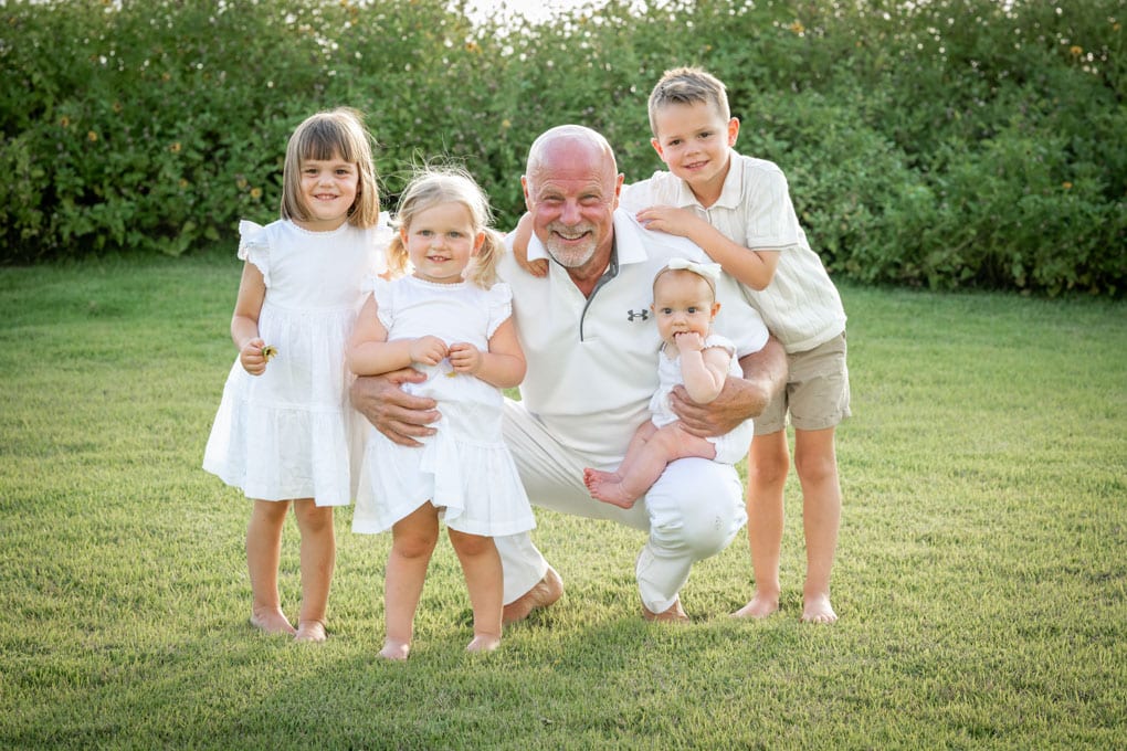 An older man kneels on grass, smiling and surrounded by four barefoot children dressed in white—posing together in a sunny garden, captured with the warmth and artistry of a Naples beach portrait photographer.