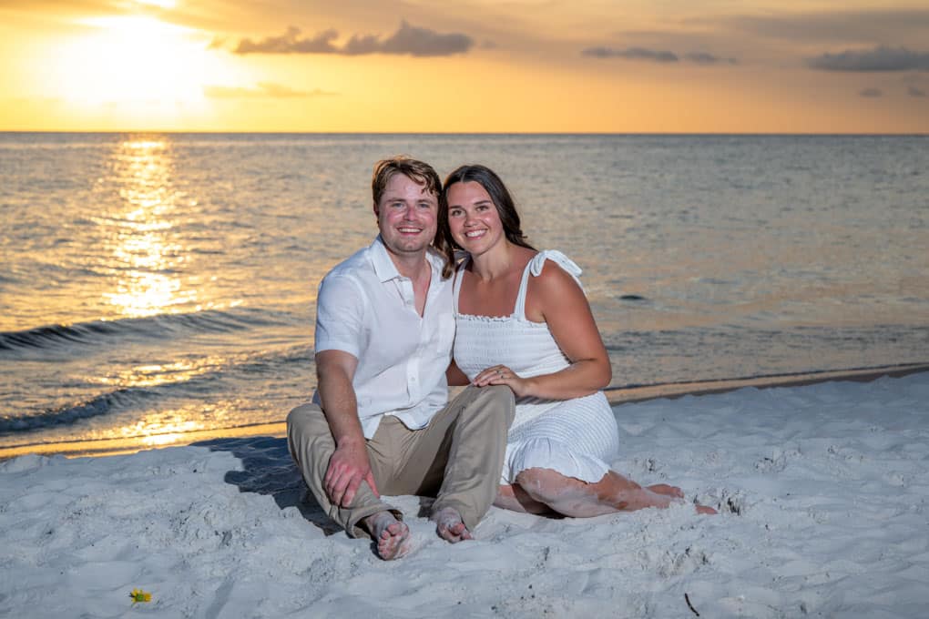 A smiling couple sits close together on sandy beach at sunset, facing the camera. Captured by a Naples beach portrait photographer, the ocean and a golden sky with scattered clouds create a dreamy backdrop for their summery attire.