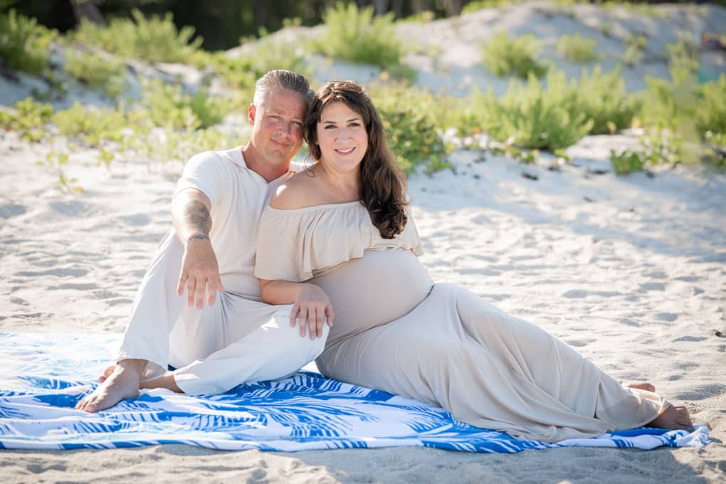 A couple sits on a blue and white blanket at the beach, captured by a Naples beach portrait photographer. The woman, in a light off-the-shoulder dress, is pregnant and smiling beside her partner, with greenery and sand in the background.
