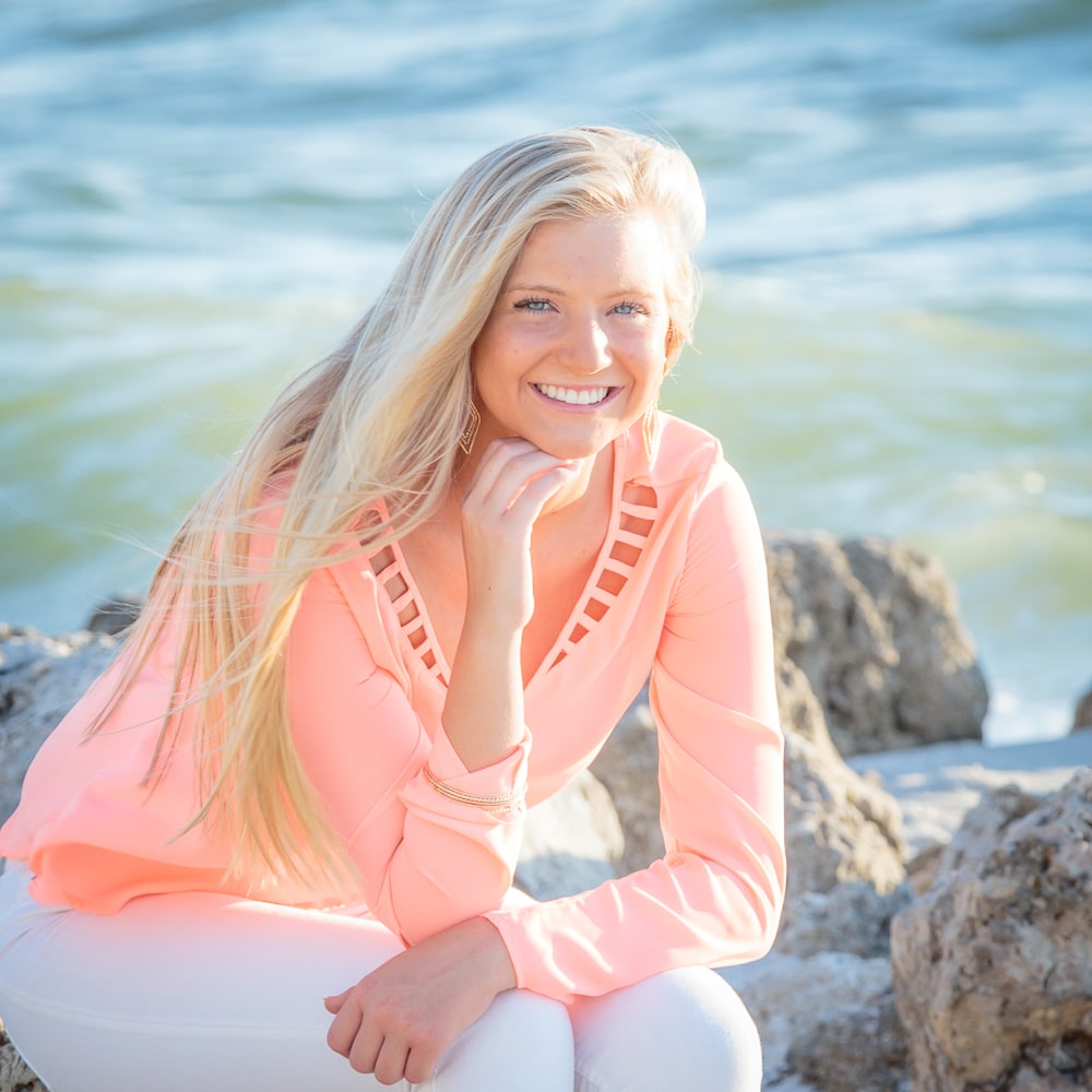 A smiling woman with long blonde hair, wearing a light pink blouse and white pants, sits on rocks by the seaside with gentle waves in the background.