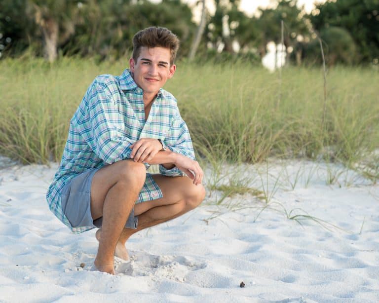 A young man in a plaid shirt and shorts squats barefoot on white sand at a beach, with tall green grass and trees in the background. He is smiling at the camera.
