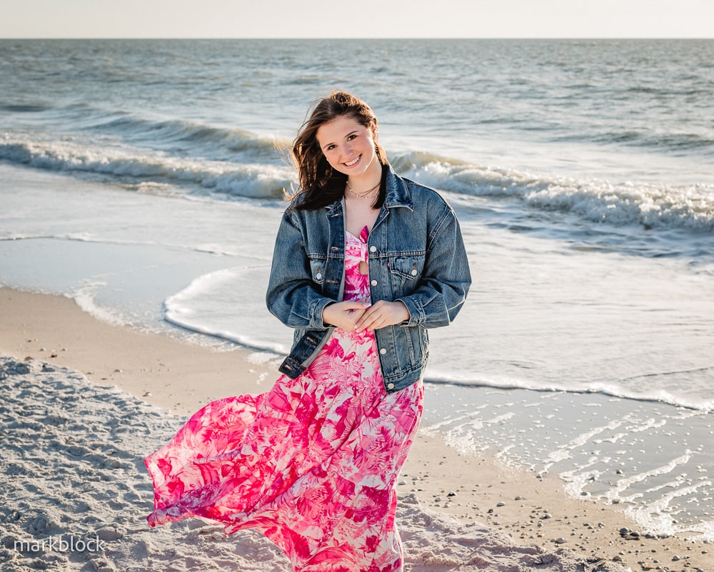 A young woman in a pink floral dress and denim jacket stands smiling on a sandy beach with gentle waves in the background, sunlight illuminating her and the ocean.