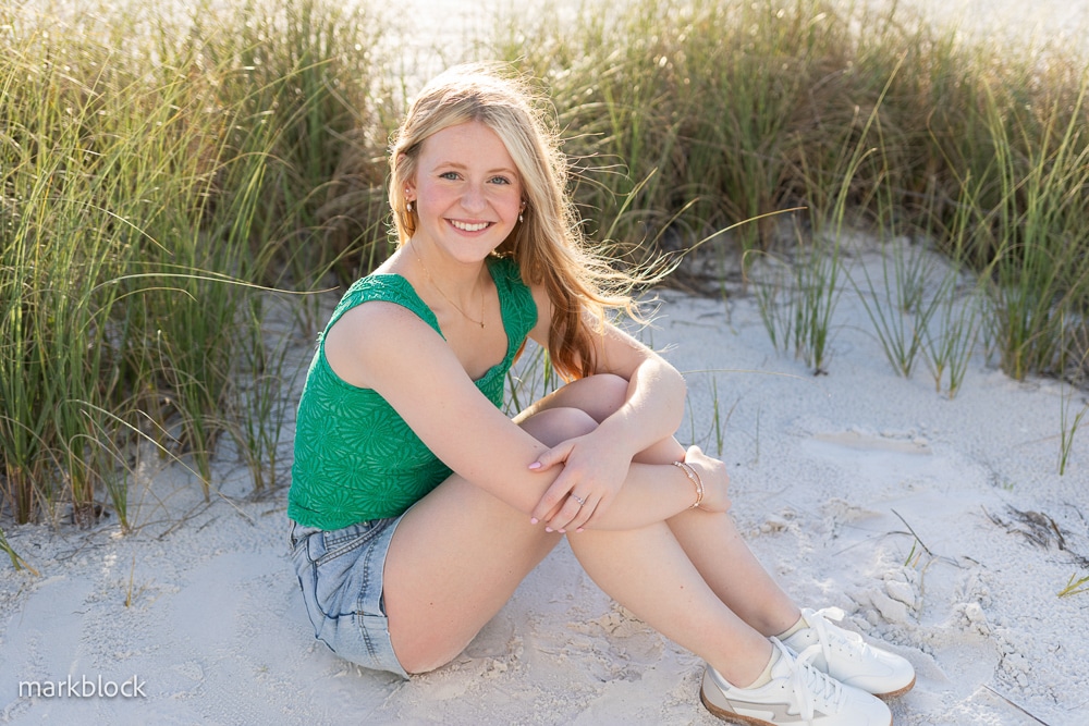 A young woman with long blonde hair, wearing a green sleeveless top, denim shorts, and white sneakers, sits smiling on sandy ground with tall grass in the background on a sunny day.