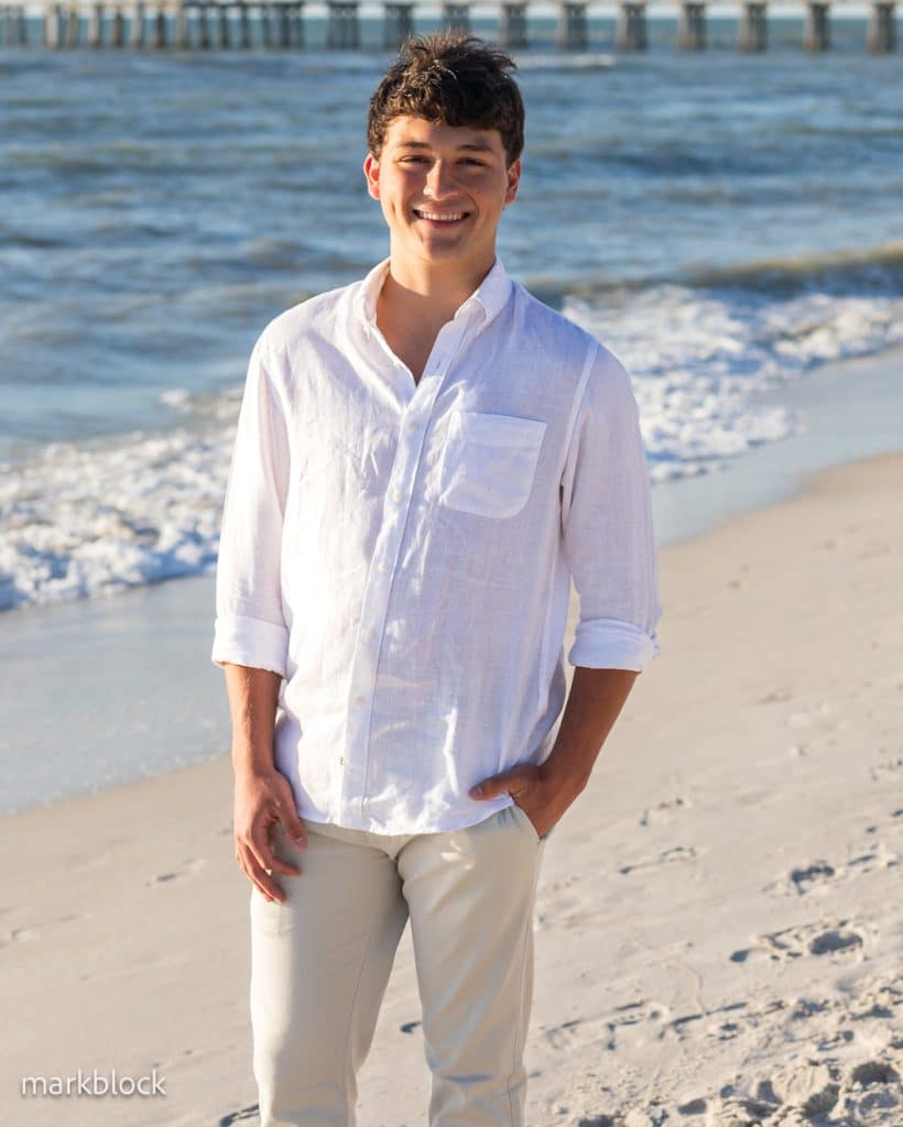 A young man wearing a white shirt and light pants smiles while standing on a sandy beach with waves and a pier in the background.
