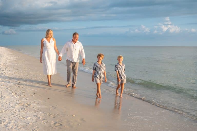 A family of four, two adults and two children, walk barefoot along a sandy beach near the ocean, enjoying a sunny day with blue skies and light clouds—capturing moments like these is why knowing how to choose a beach photographer matters for preserving special memories.