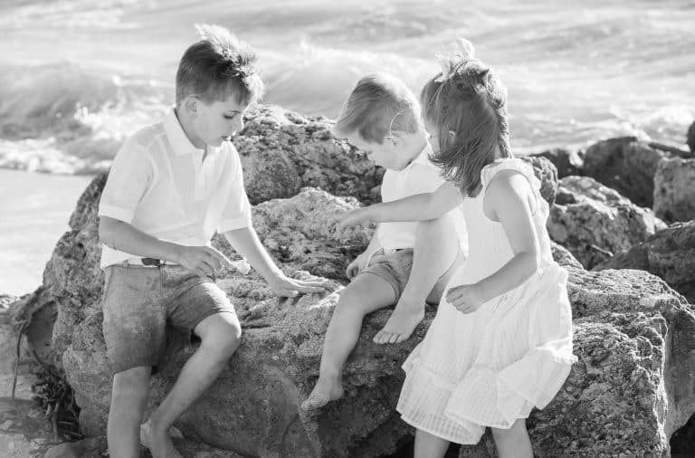 Three young children, two boys and one girl, sit and play on large rocks by the seaside during a windy day—perfect inspiration for your Marco Island portrait session guide. Waves crash in the background as they laugh in their light summer outfits.