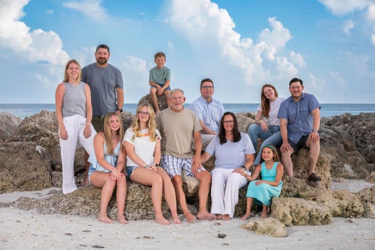 A group of eleven people, including adults and children, pose together on rocks at a beach near Marco Island and Naples, with blue sky and clouds in the background. Everyone is smiling and dressed in casual summer clothes.