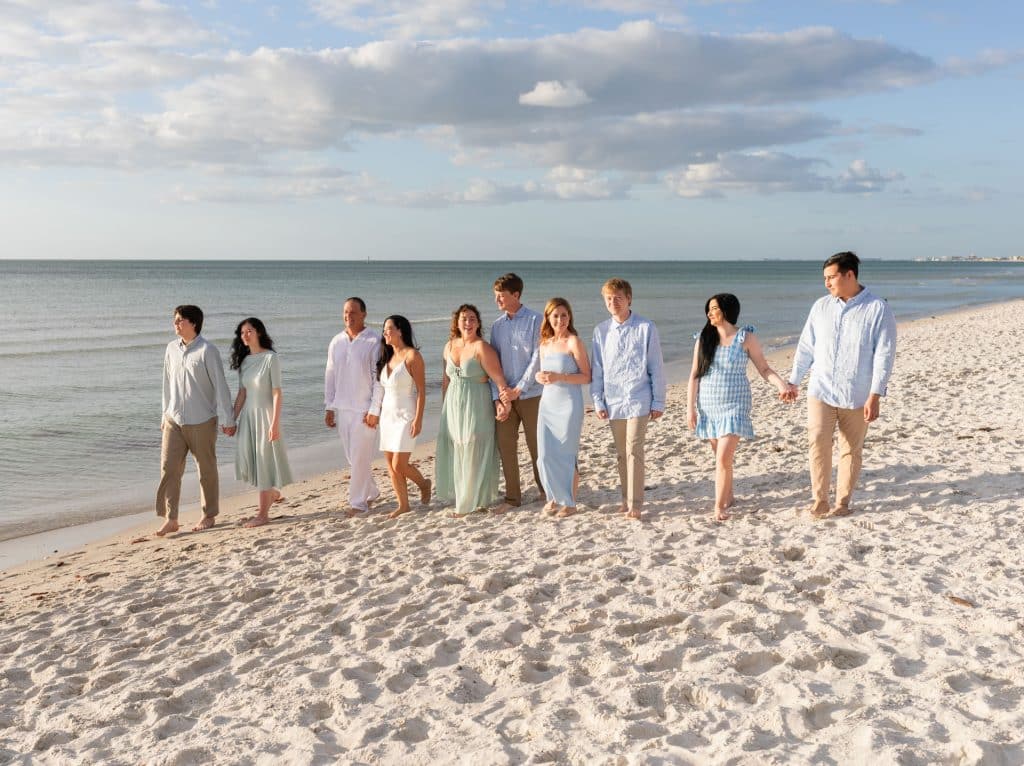 Nine people walk together along a sandy beach by the ocean, dressed in light-colored, coordinated outfits, under a partly cloudy sky. They appear happy and relaxed, enjoying the sunny day.