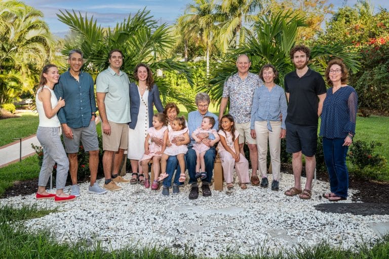 Thirteen adults and six young children pose together outdoors in a lush, green garden with palm trees, all smiling at the camera.