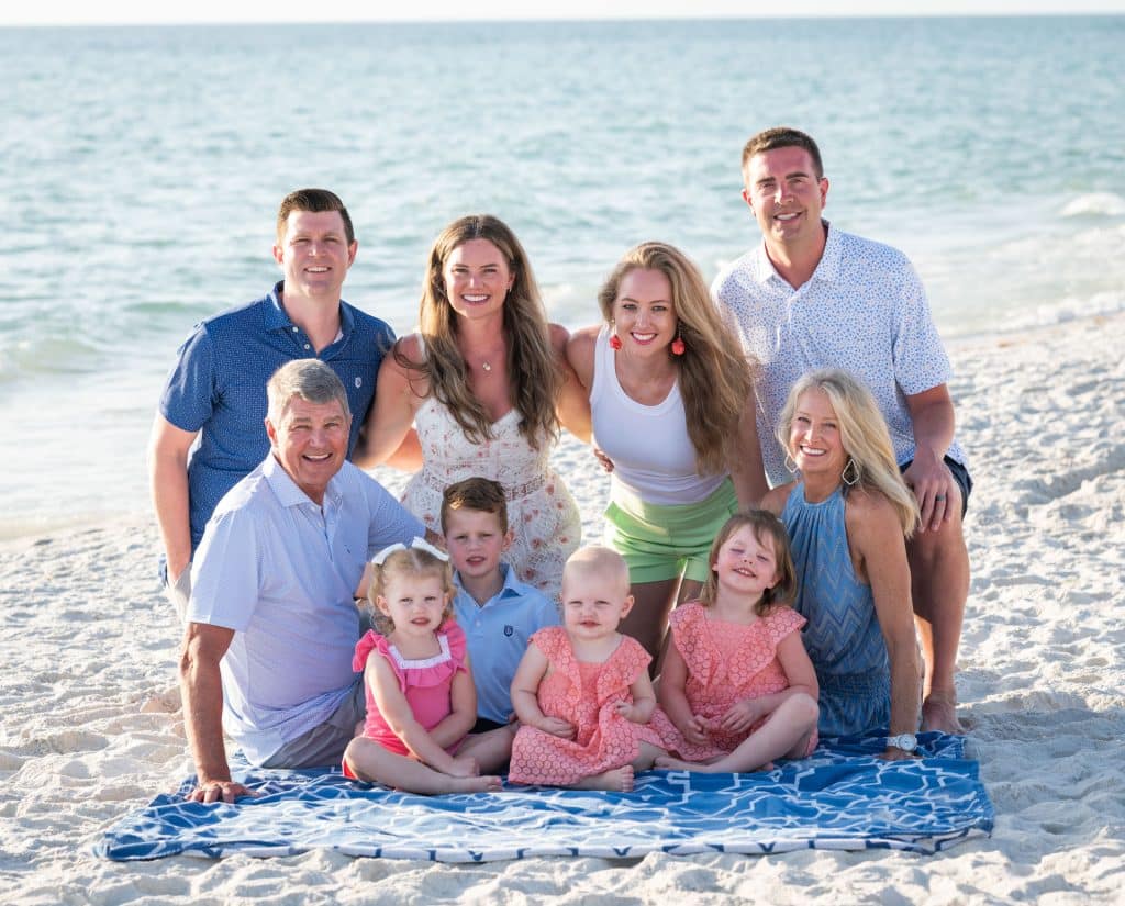 Three men, two women, and four small children sit and kneel together on a sandy beach by the ocean in Naples, FL, smiling at the camera. The group is on a blue blanket, with calm water and a clear sky in the background.