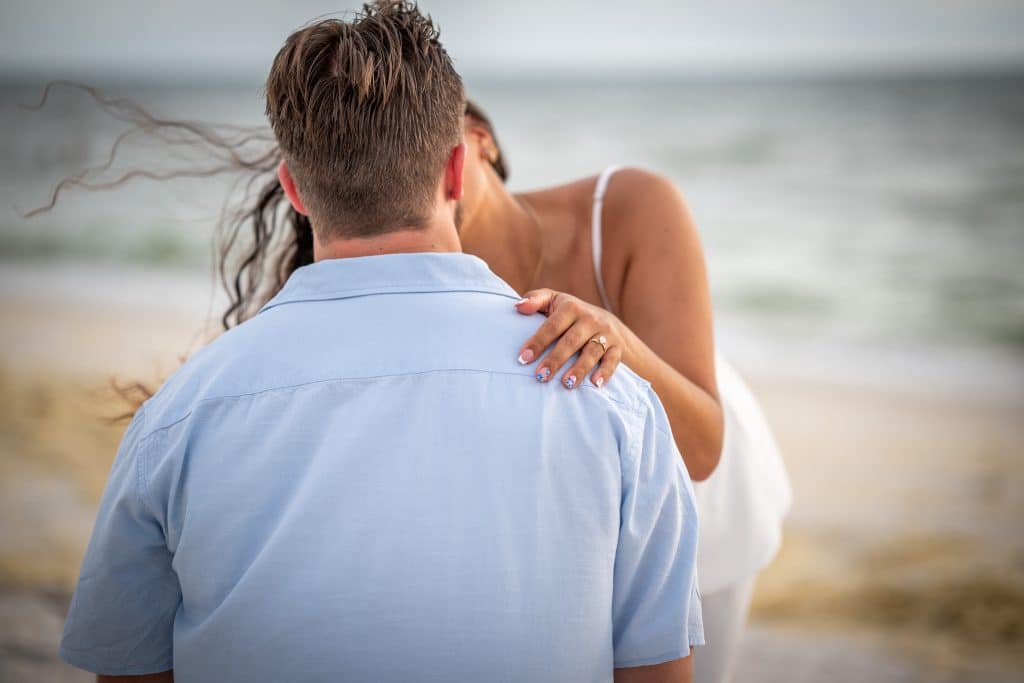 A woman hugs and kisses a man from behind on a beach. Her hand, with a ring, rests on his shoulder. Both are dressed in light clothing, and the ocean is blurred in the background.