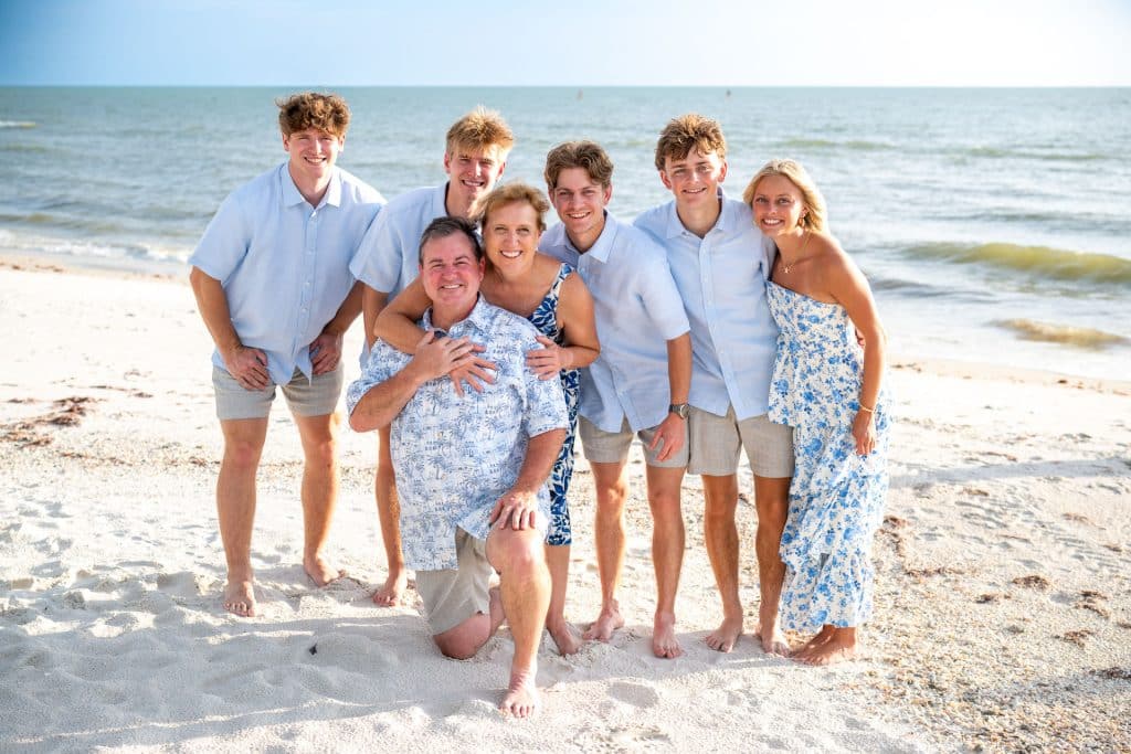 A family of seven poses and smiles on a sandy beach by the ocean, dressed in coordinating blue and white outfits under bright sunlight.