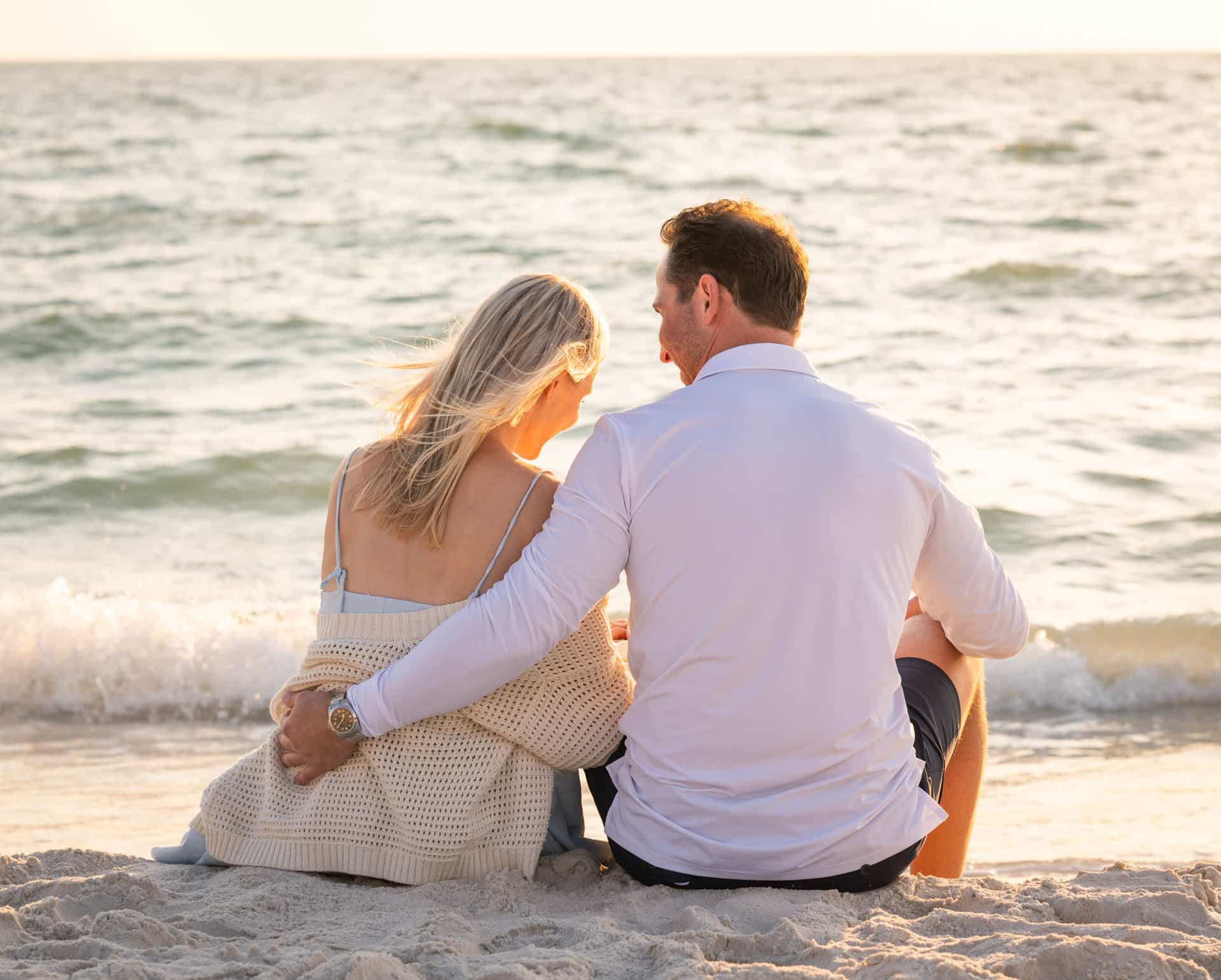 A couple sits closely together on the beach in Naples and Marco Island, facing the ocean at sunset. The woman wears a light sweater, and the man has his arm around her. Waves gently approach the shore in the background.
